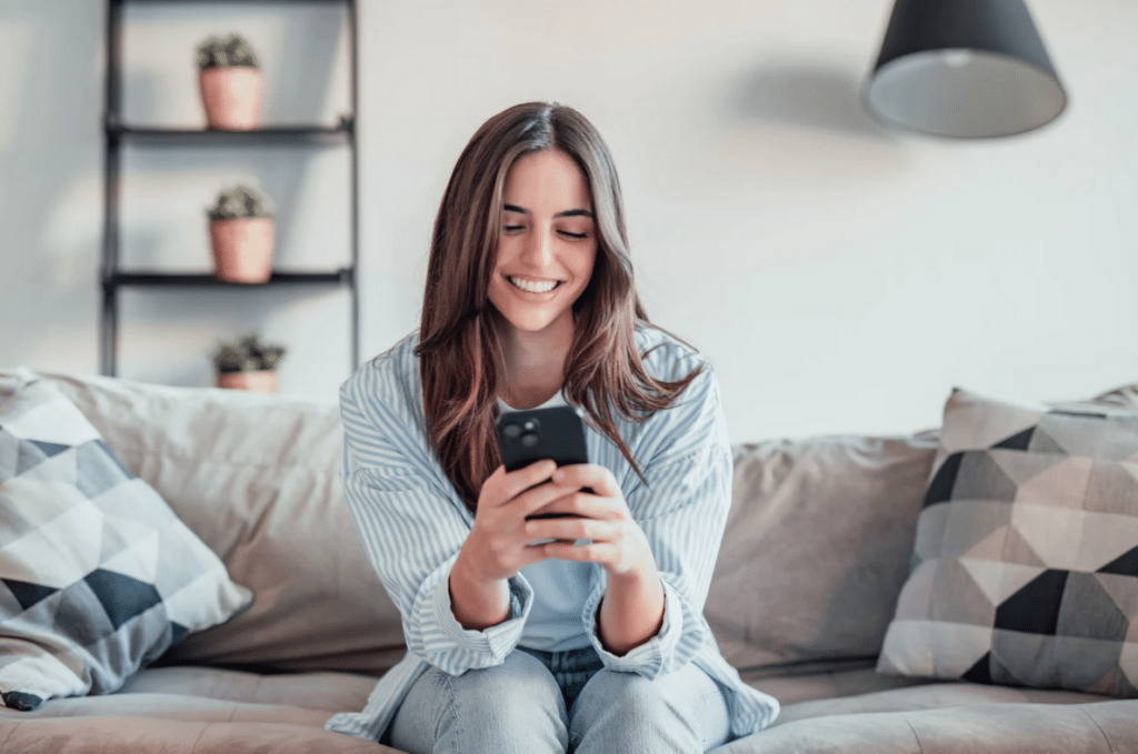 A young woman sits on a couch, smiling while looking at her smartphone. There are decorative pillows and potted plants in the background.